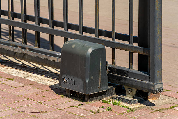 An electric gate motor is placed on the ground beside a metal fence. The gate is in an open position. The setting is outdoors during daylight hours.