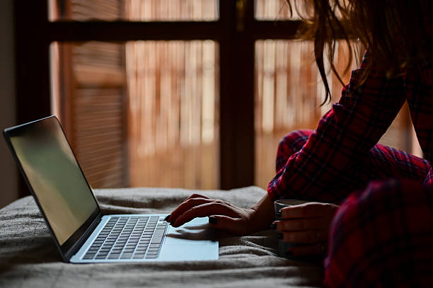 Woman typing on laptop in bed wearing pajamas, enjoying a warm cup of coffee Woman typing on laptop in bed wearing pajamas, enjoying a warm cup of coffee
