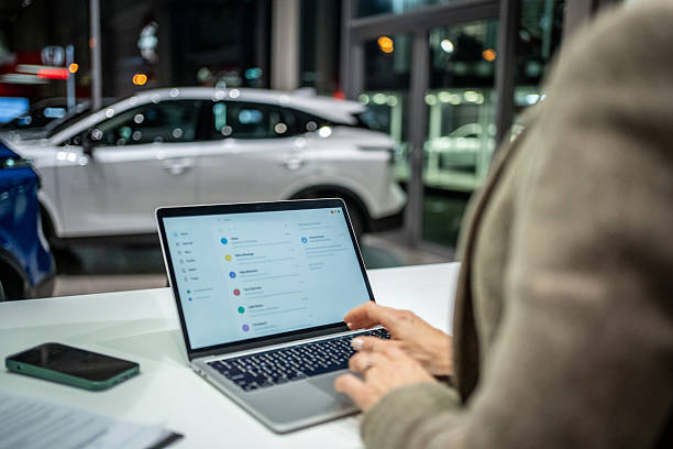 Woman focusing on her laptop screen, typing on the keyboard while sitting at a desk in a modern car showroom environment, staying connected and managing business